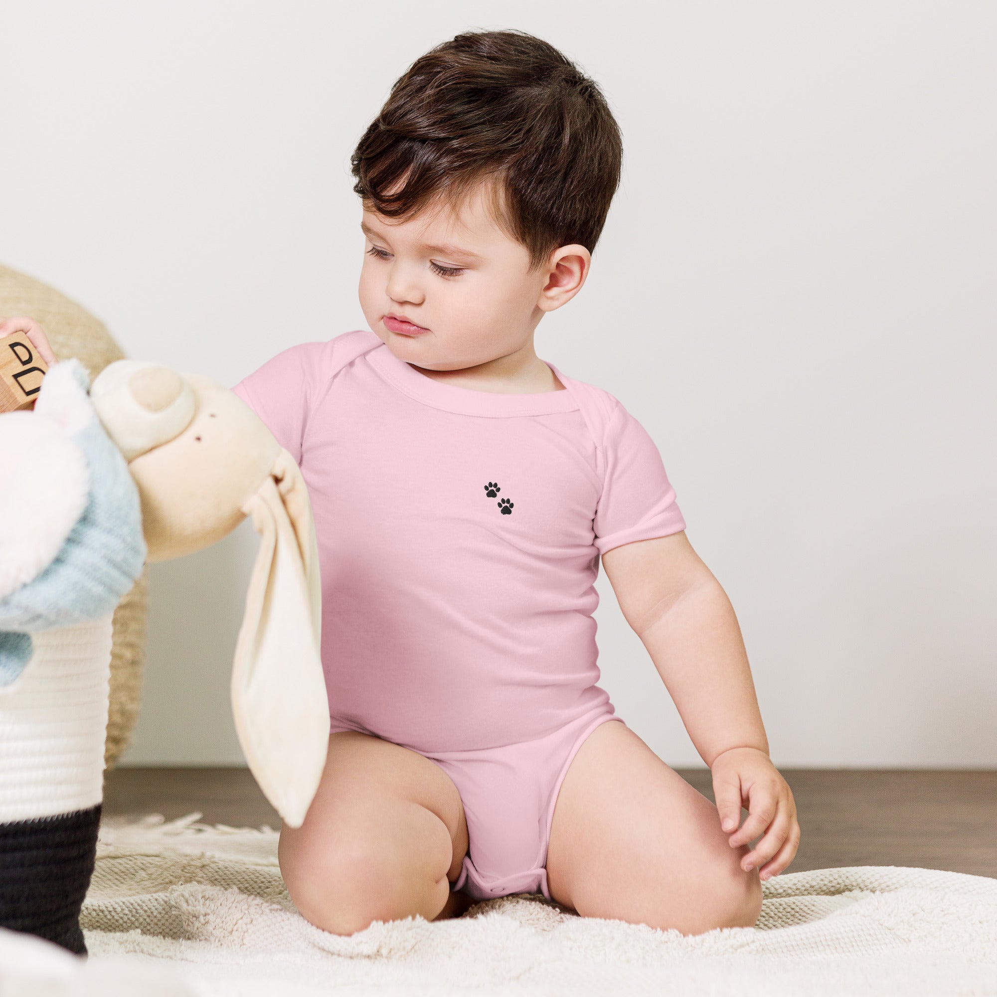Child wearing a pink onesie with a small logo, sitting on a carpeted floor.