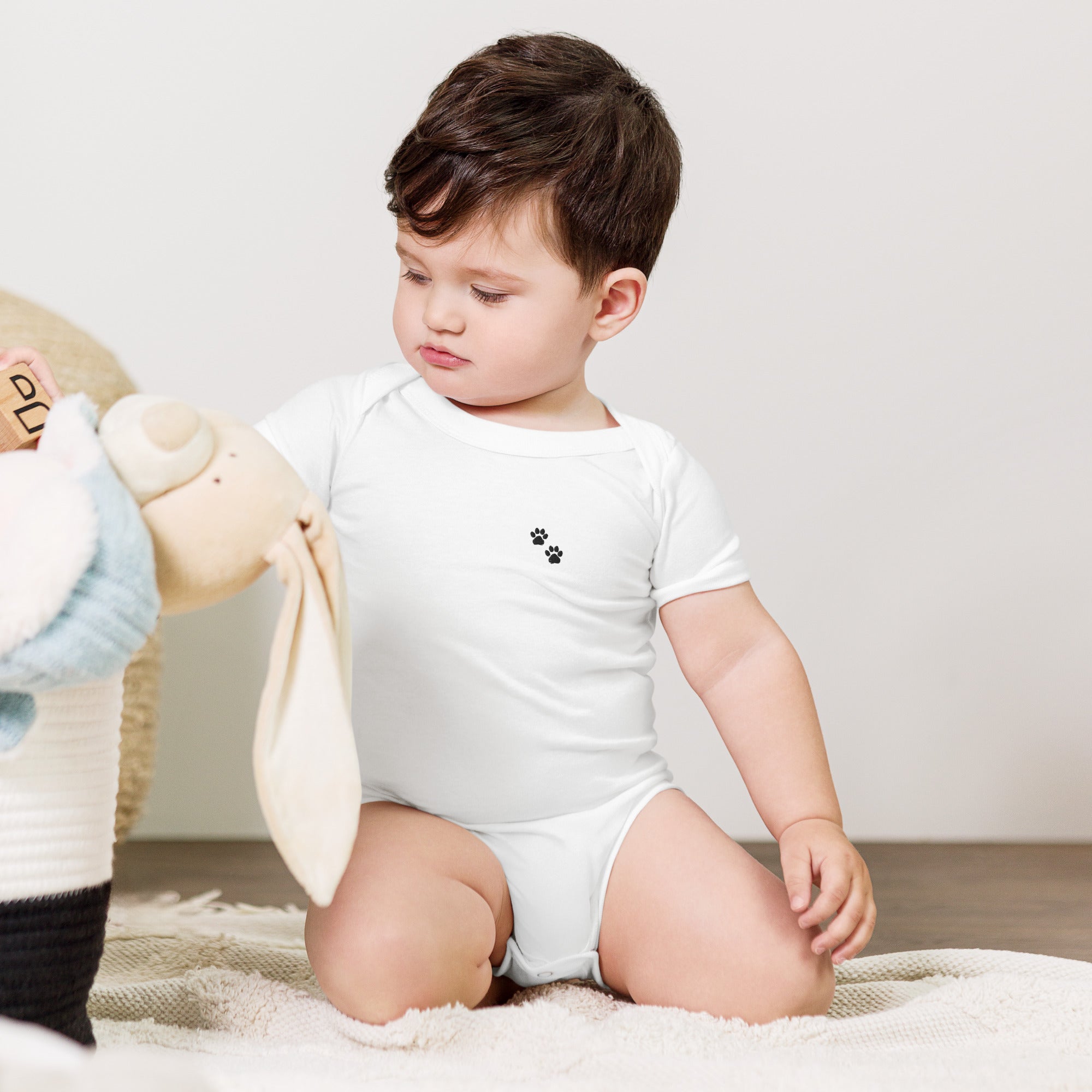 Child wearing a white onesie with a small design, sitting on a carpeted floor.