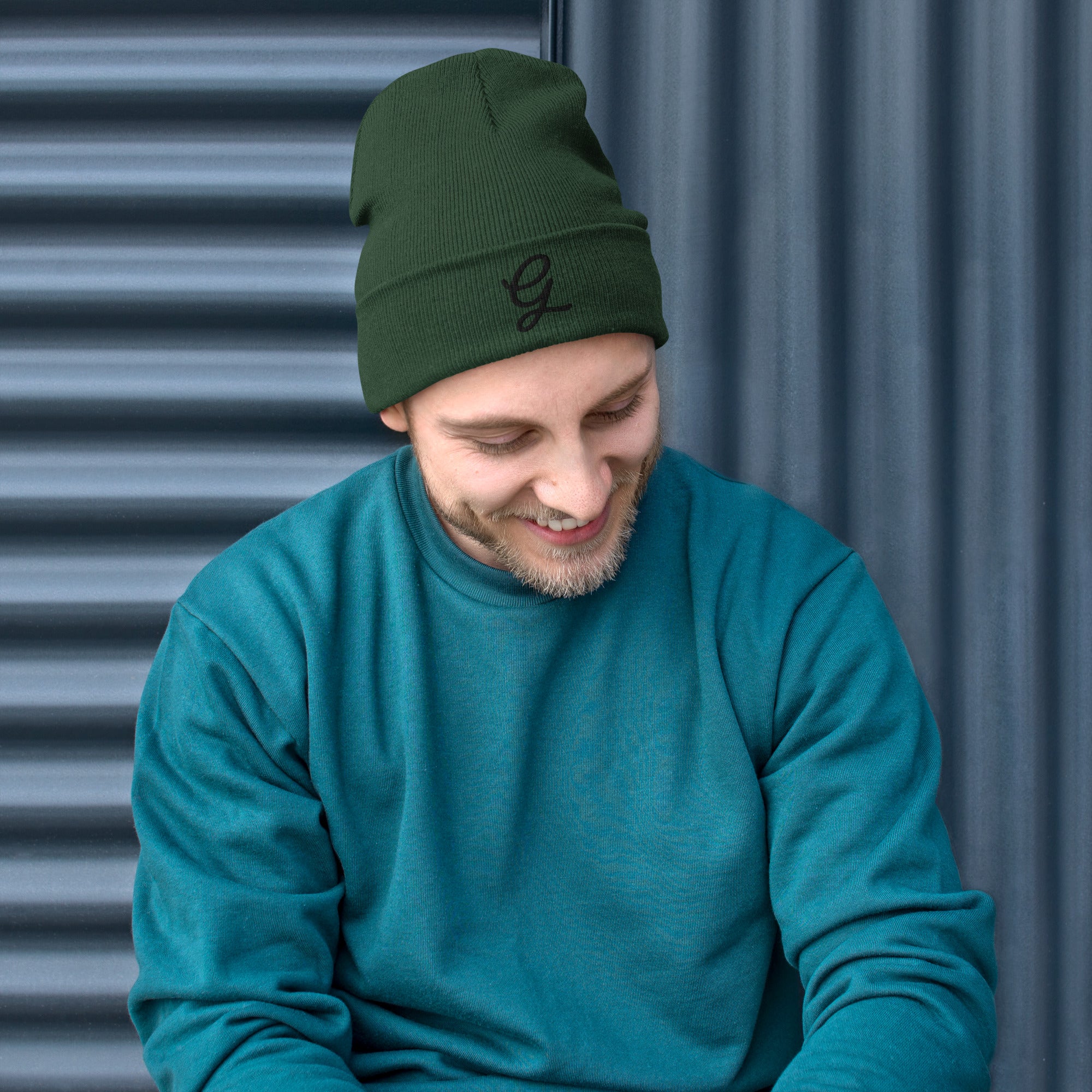 Man wearing a granpaw golf green beanie and teal sweater sitting against a corrugated metal background