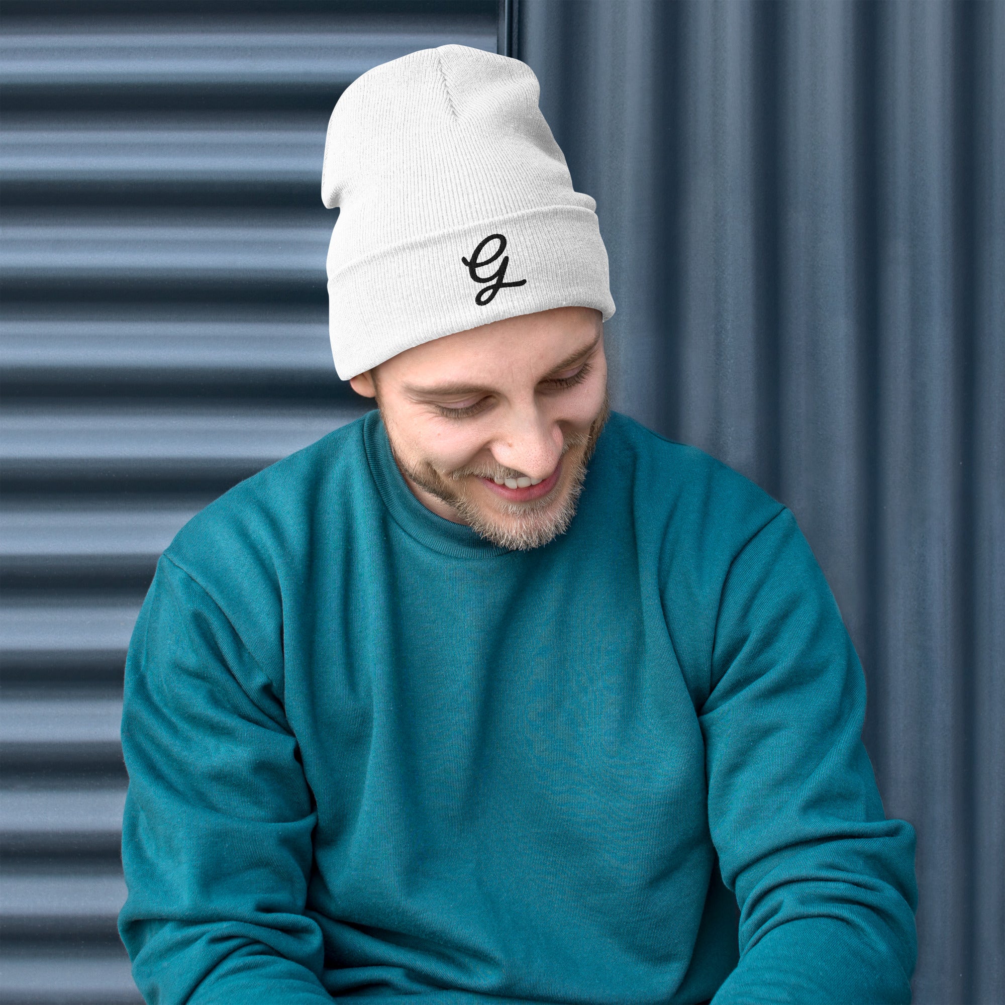 Man wearing a white beanie with a granpaw golf logo and a teal sweater against a corrugated metal background.