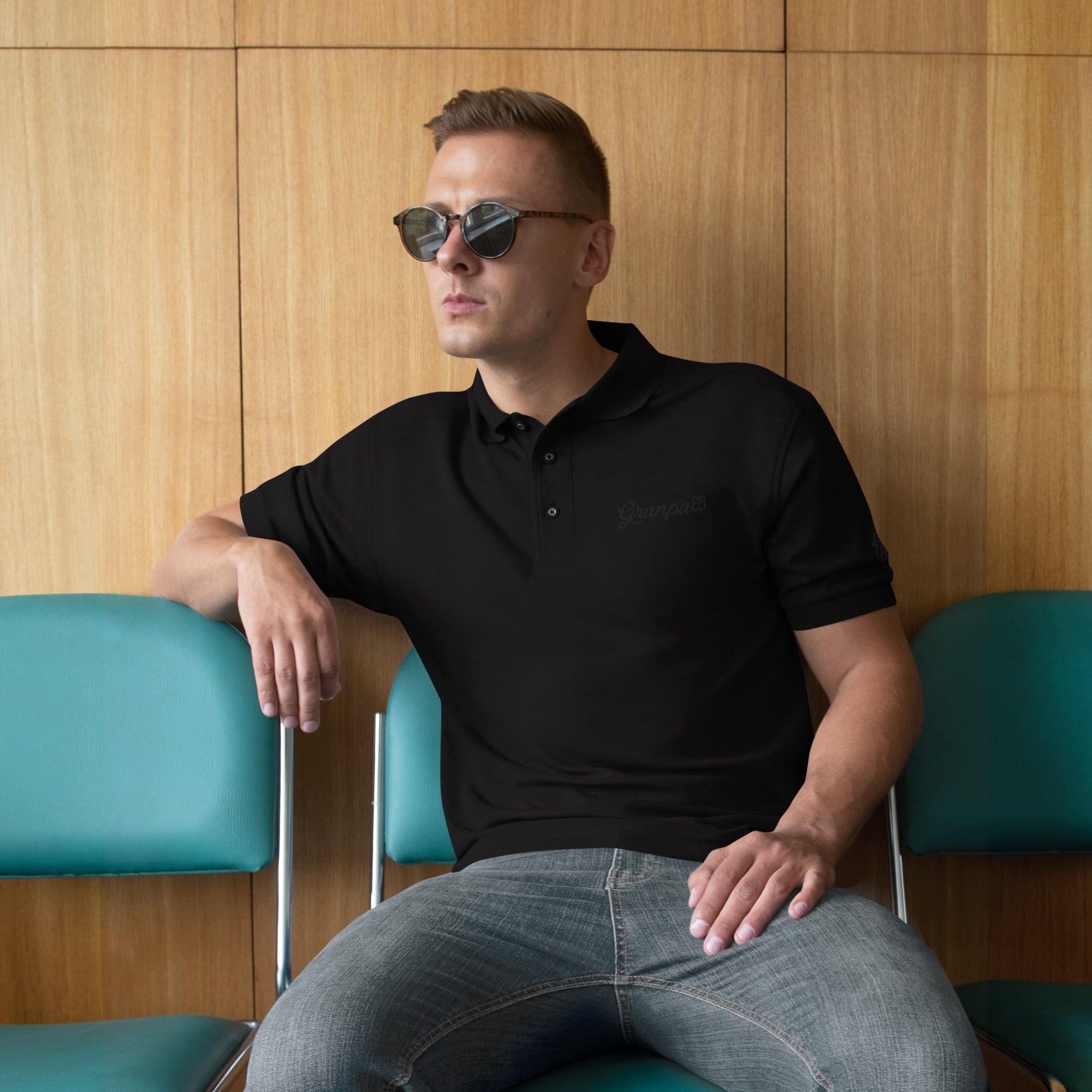 Man wearing a black polo shirt with sunglasses sitting against a wooden wall.