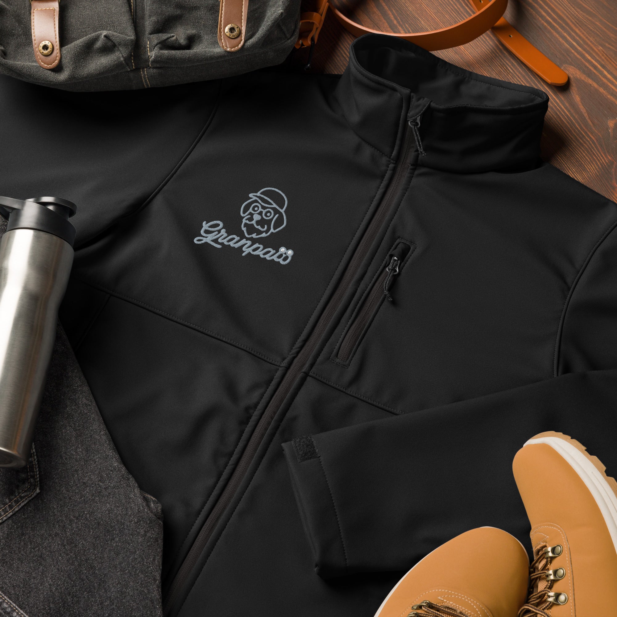 Black jacket with a brand logo on a wooden surface with backpack, shoes, and tumbler.