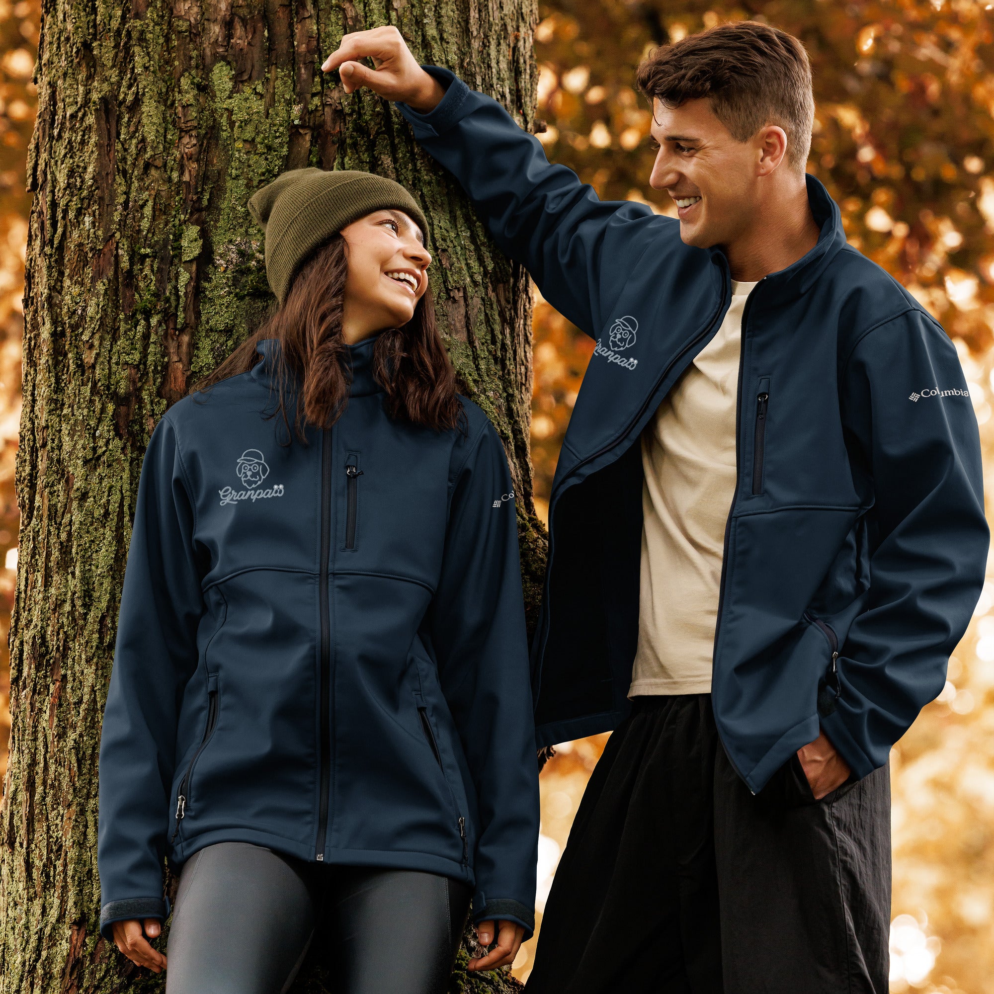 Two people wearing Columbia jackets standing next to a tree in an autumn setting.