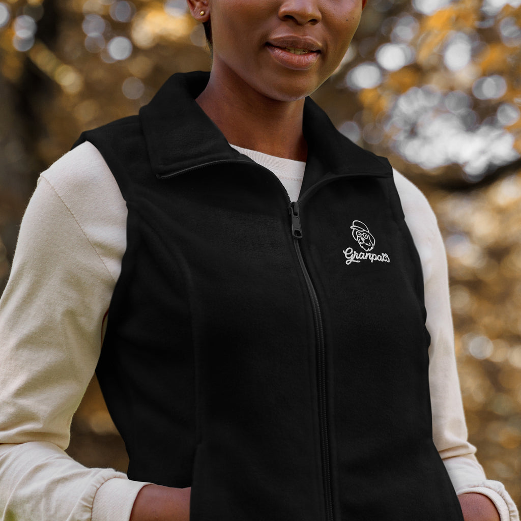 Person wearing a black vest with a logo, standing outdoors with blurred foliage in the background