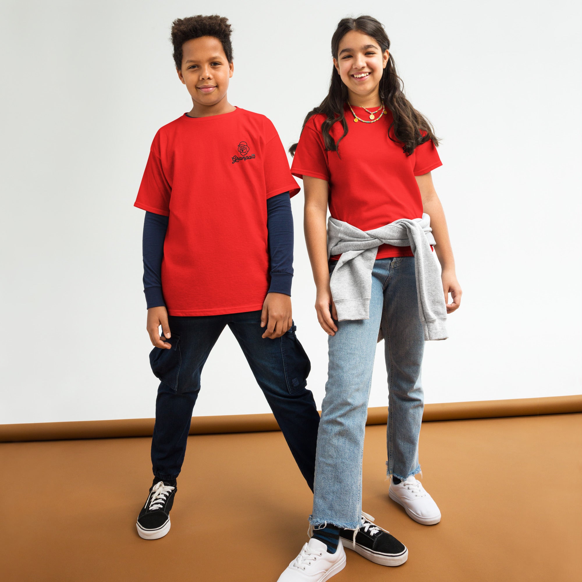 Two children wearing red shirts standing on a brown floor with a white wall background