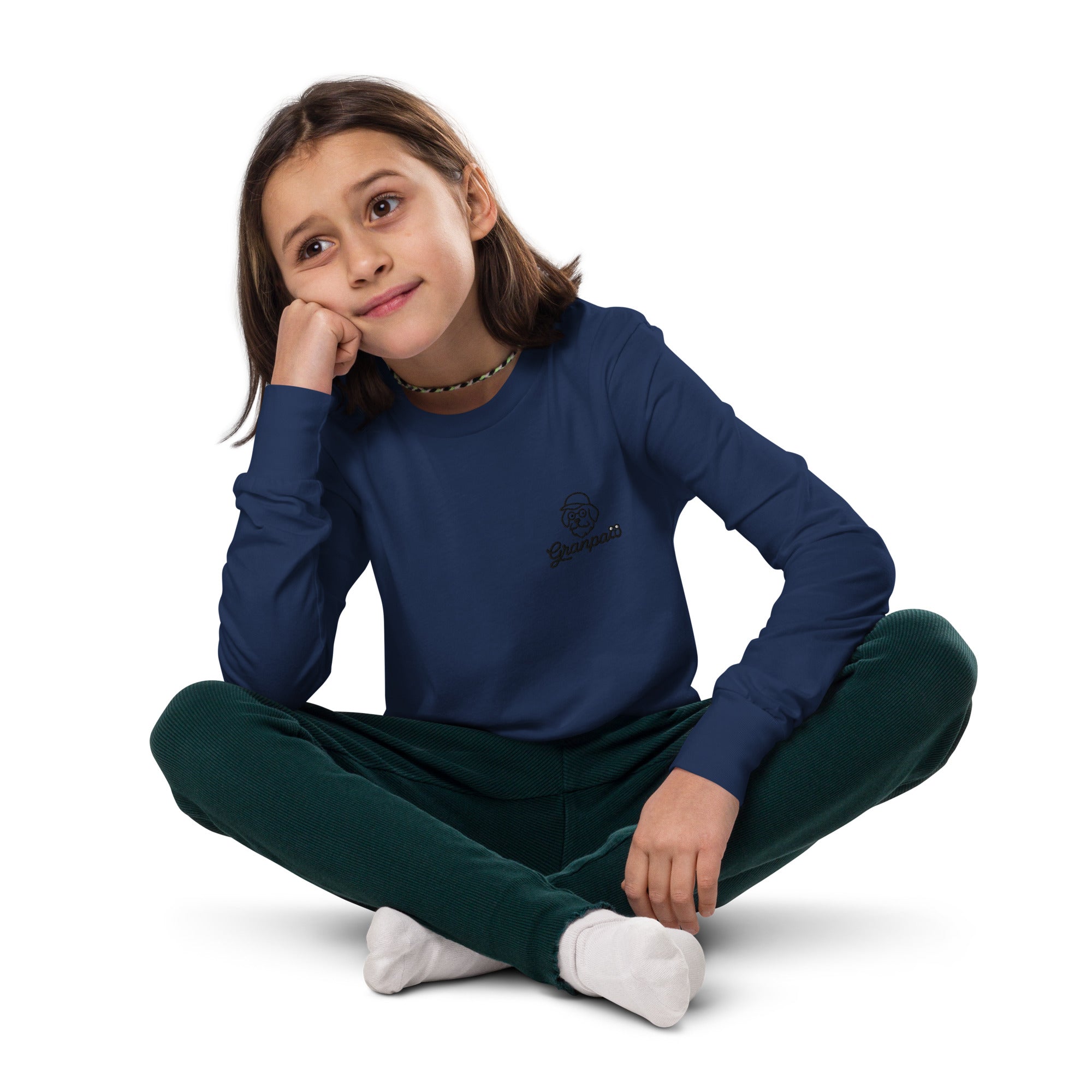 Young girl wearing a navy blue long-sleeve shirt and green pants sitting on a white background